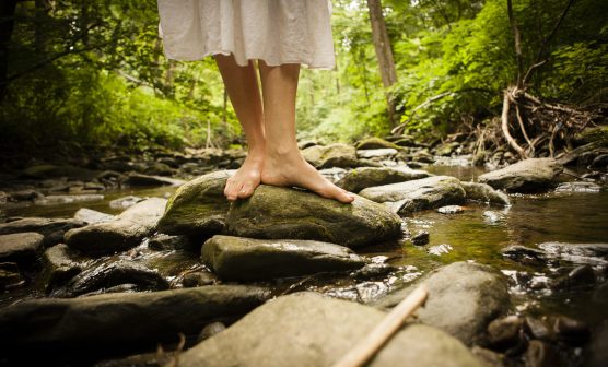 Bare feet of mid adult woman wearing white dress balancing on rock in riverbed