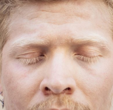 Close up portrait of the facial features of a Latino albino man. His long white lashes shield his blue eyes from the sunlight.